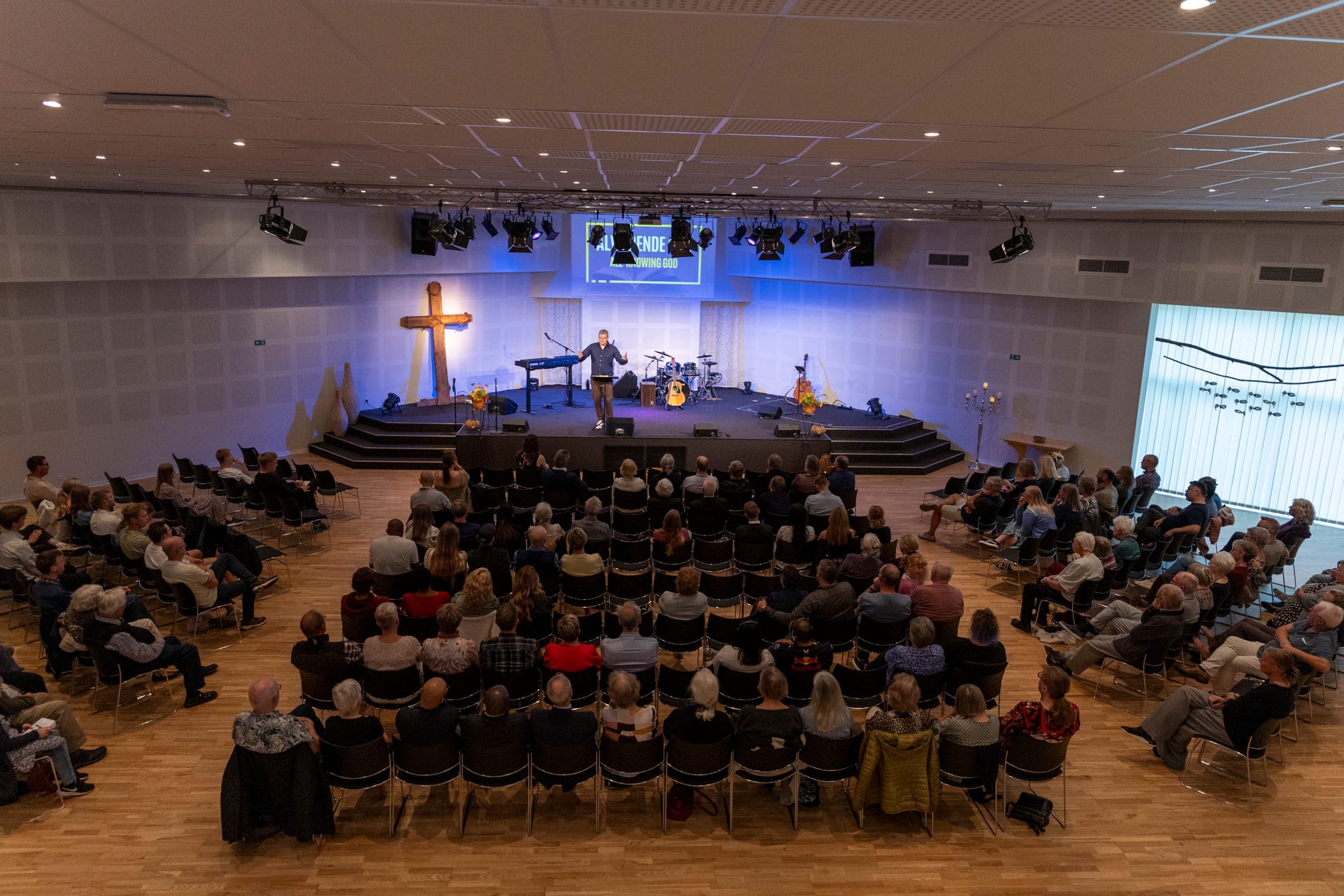 La sala de la iglesia llena de personas durante el culto, con el escenario y la cruz de madera al frente