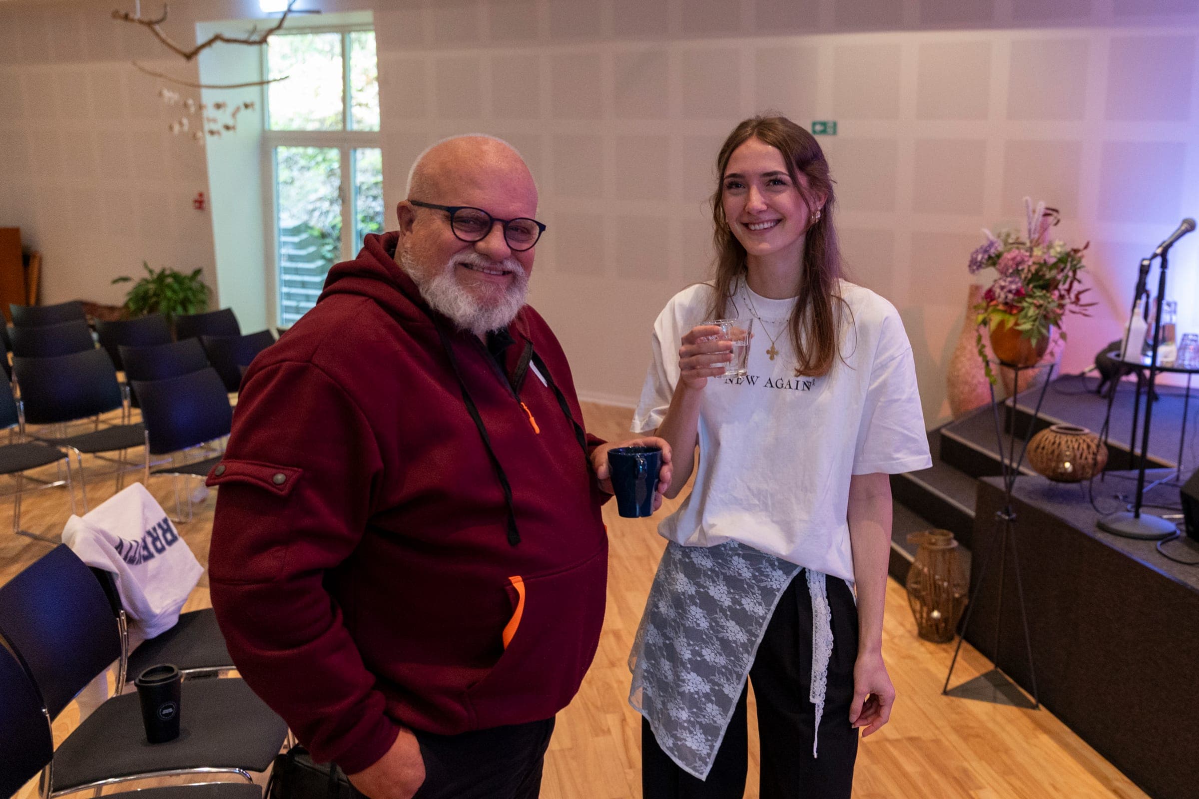Zwei Menschen aus der Kirche lächeln einladend mit Kaffee in der Hand im Kirchensaal